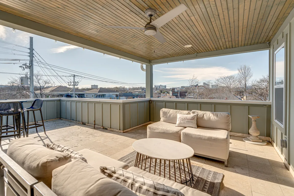 A sophisticated outdoor seating area on a rooftop deck, showcasing comfortable lounge furniture under a covered section, a ceiling fan, and a breathtaking view of the Nashville skyline. The city lights twinkle in the background, creating an inviting setting for evening relaxation or socializing.