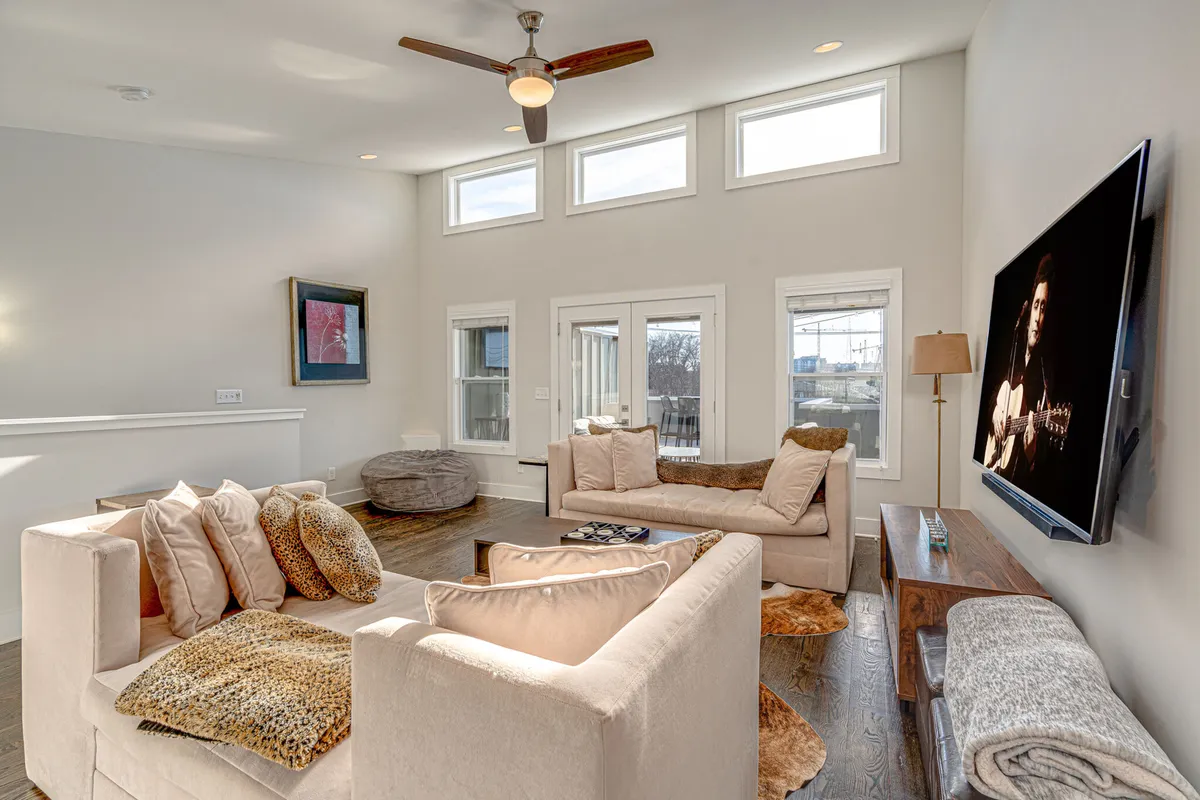 A chic, cozy secondary living area featuring soft beige sofas arranged around a large flat-screen TV, with expansive windows flooding the room with natural light. An elegant art piece decorates the wall, complementing the modern, welcoming ambiance. The setup is perfect for relaxing, socializing, or hosting lively gatherings.