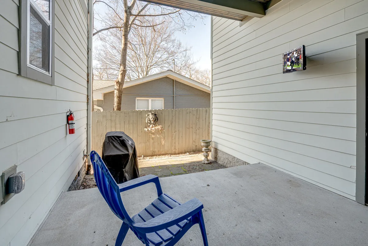 A fenced backyard with a blue outdoor chair, TV mounted on the wall, grill, and green space—suggesting both relaxation and entertainment options for guests and their pets.