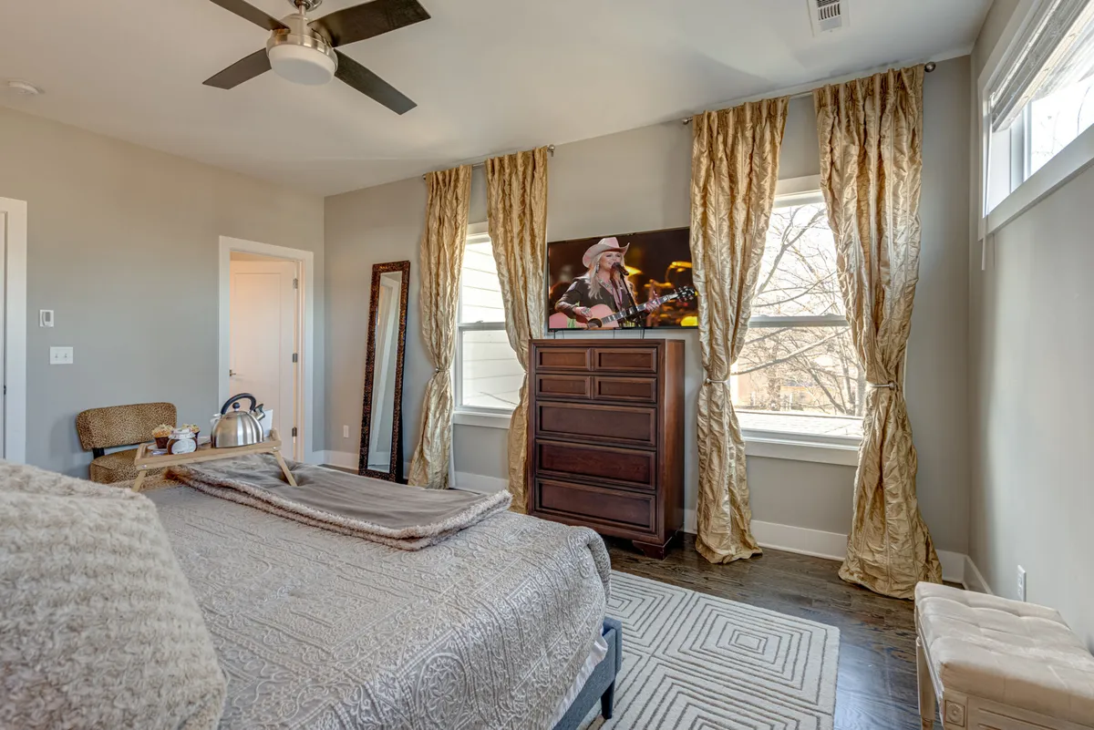 A queen-sized bed with a decorative blanket faces a television atop a wooden dresser. Large windows with curtains admit ample daylight, and a mirror adds a sense of space, creating a restful, inviting bedroom setting.