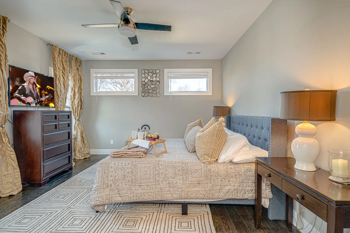 A king-sized bed is topped with decorative pillows, flanked by nightstands and lamps. A television displays a musician, and soft drapery filters natural light, creating a serene and comfortable environment ideal for unwinding.