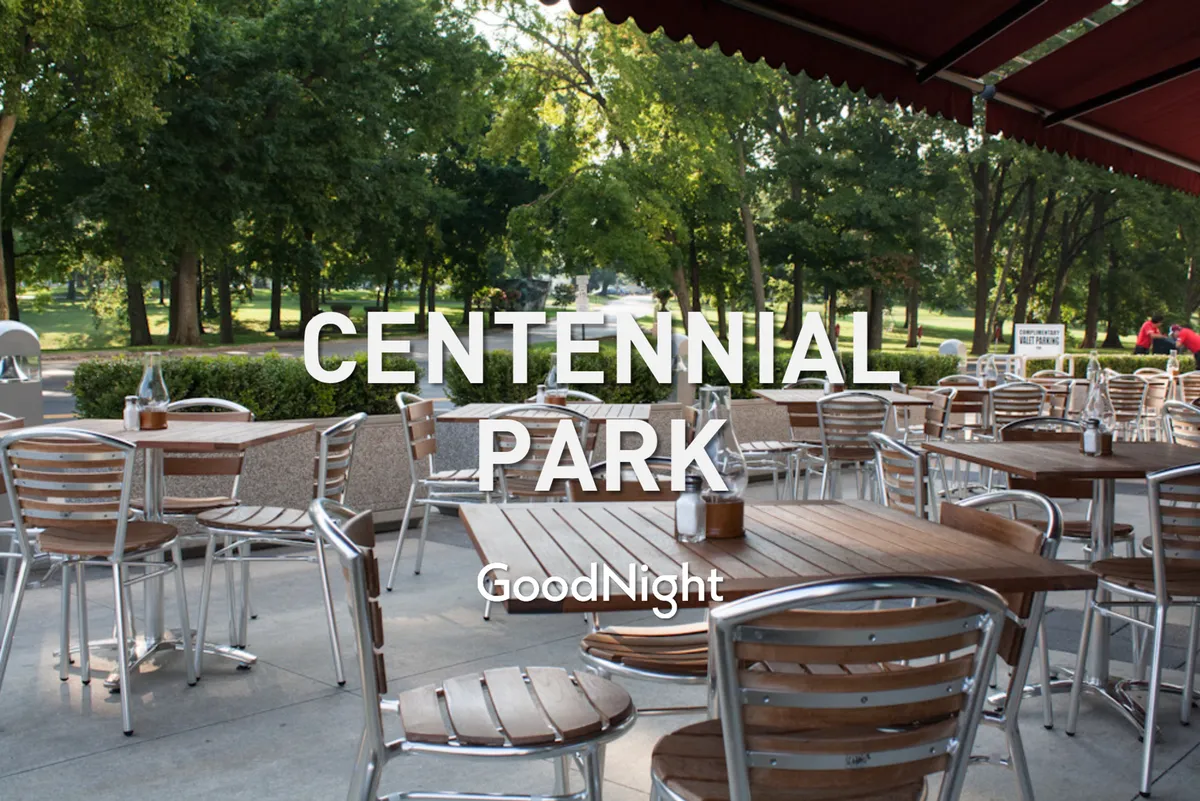 An inviting outdoor dining area with wooden tables, metal chairs, and lush green trees surrounding the space. There is a walkway leading into a park, suggesting easy access to green space for walks and relaxation with pets.